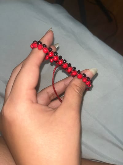 A person's hand, characterized by long, natural-toned fingernails, is centrally featured, holding a beaded bracelet. The bracelet is crafted from small, glossy beads in alternating red and black colors, strung together on a red cord. The design appears to be a macrame style, with the beads secured by knots along the cord, and loose red cord ends are visible near the palm.
The activity depicted is the handling or examination of the bracelet. The hand is gently cupped, showcasing the jewelry.
The scene is an indoor setting, likely a private space such as a bedroom. The background consists of soft, light-colored fabric, possibly bedsheets or a blanket, which is slightly wrinkled. The lighting is dim and diffused, suggesting either evening hours or an interior lit by artificial light. Shadows are present, particularly in the upper left portion of the image. There are no discernible emotions conveyed by the hand. No text is visible in the image.