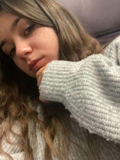 A close-up, angled portrait features a young female individual from the mid-chest upwards, situated slightly to the right of the frame, looking directly towards the camera. Her head is tilted, and her right hand supports her chin, with her fingers curled. She wears a light grey, heathered knit sweater with a prominent horizontal rib pattern on the sleeve. A metallic ring is visible on her right ring finger, partially obscured by the sweater cuff. She has long, wavy, medium-brown hair that falls over her shoulders. Her eyes are dark, and her facial expression is neutral. The background is dark and indistinct, offering no discernible environmental features. The exact location within Schwindratzheim, France, cannot be determined from the visual information presented in the image.