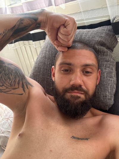 A shirtless man with a full beard and short hair is lying on his back on a pillow. He has tattoos on his left arm and a small tattoo on his chest. His right fist is resting on his forehead. The background shows a window with white curtains and a black and white striped fabric. The setting appears to be an indoor space, possibly a bedroom or living room. The image is taken from a close-up perspective, focusing on the man's face and upper body. There is no visible indication of the city of Stadtroda, Germany in the image.