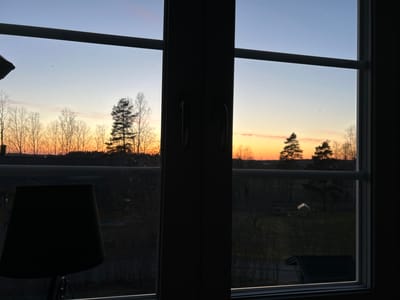 A view from a window in a dwelling in Gränsta, Sweden, shows a twilight or dawn landscape. The window, featuring multiple panes and dark frames, partially obscures the view. Outside, a mix of deciduous and coniferous trees line the horizon, silhouetted against a gradient sky with hues of blue, orange, and yellow. Below the horizon, rooftops of nearby buildings are faintly visible. The foreground includes a lamp with a dark shade partially visible on the left and a blurry outdoor scene with sparse vegetation. The overall ambiance suggests dusk or dawn in a suburban or rural setting.