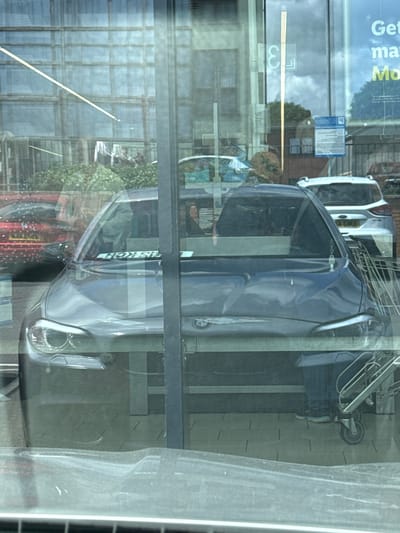 A mid-shot of a dark grey BMW car seen from the front and slightly from the side through a glass surface which reflects trees, buildings and a blue sign. The car is parked behind a metal shopping trolley. The car has its headlights on and its bonnet is visible. A number plate on the car's windscreen reads 'HON SIL'. Behind the BMW, a silver SUV is visible with a visible number plate 'EX13 ENE'. The scene appears to be a car park, possibly at a supermarket in Kingswood, United Kingdom. A person wearing blue jeans and white shoes is visible standing next to the shopping trolley on the right side of the frame.