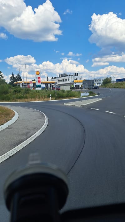 The image depicts a gas station and an autohof complex located in Haiger, Germany. A Shell gas station with visible price displays and signage is prominent on the left. Adjacent to it, an "AUTOHOF" building with a yellow "X" logo is visible. The scene is set under a bright blue sky with scattered white clouds. A curved asphalt road, likely a highway exit or entrance, dominates the foreground. The road has white lane markings and a concrete barrier. The image appears to be taken from inside a vehicle, possibly from a dashboard mount, given the blurry foreground element which resembles a camera mount. There are no visible people in the image, and the focus is on the infrastructure of the location.