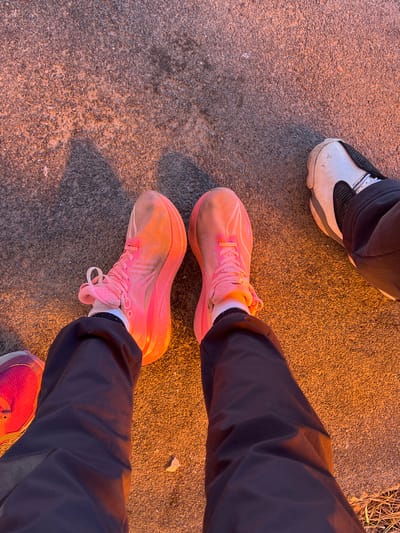The image shows a street surface with three pairs of shoes visible. One pair is white and gray sneakers, worn by a person whose legs are partially visible at the top of the image. Another person stands wearing pink running shoes and dark pants. A small portion of the person's other shoe is visible in the bottom right of the frame. The street surface is textured. The lighting suggests it might be twilight or artificial lighting. Gory, Russia, is not discernible from the image.