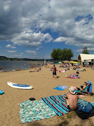 This is a ground-level, eye-level photograph taken at the Radslav Lake beach in the Czech Republic on a sunny day with scattered clouds. The sandy beach is populated with numerous people engaged in various leisure activities. In the foreground, two paddleboards are visible, one white and blue, the other white. A person wearing a white hat and black swim trunks sits on a colorful striped blanket, facing away from the camera. To their left, a mint green blanket with a flamingo and palm leaf pattern is spread out. Further in the foreground on the right, a woman in a black swimsuit and a straw hat reclines in a blue beach chair, her back to the camera.
In the mid-ground, a man in a maroon t-shirt and blue shorts walks across the sand. Several other individuals are sunbathing or sitting on towels and blankets. Some are in swimwear, while others are partially clothed. Yellow umbrellas are set up in the background, providing shade for some beachgoers. A thatched umbrella is also visible. A white building with a yellow trim and a pink flag on top is situated on the far right of the mid-ground.
The background features a large body of water, likely Radslav Lake, with several sailboats and smaller watercraft. A rocky barrier extends into the water. The shoreline is lined with trees, and rolling hills covered in vegetation form the distant landscape under a sky filled with cumulus clouds. The overall scene depicts a busy summer day at a lake resort.
