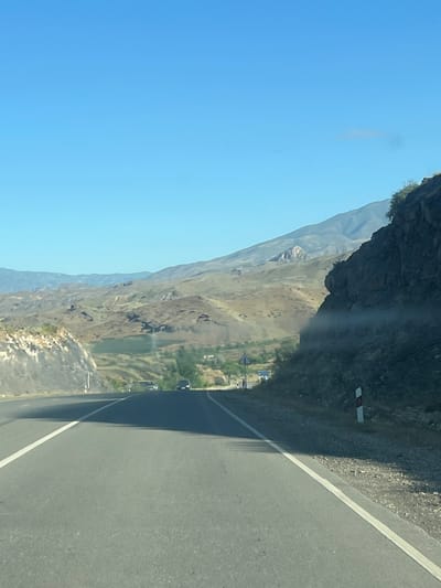 This is a vertical, eye-level, outdoor photograph taken from a vehicle moving along a paved road. The road, with a white line marking its center, dominates the right side of the frame and casts a shadow on the left. To the left of the road, a steep, rocky mountain cliff rises sharply, its dark, textured surface catching the sunlight on its upper edge.

Below the cliff, a vast valley unfolds, its slopes a tapestry of earthy tones, sparse vegetation, and some patches of green, suggesting a semi-arid environment. In the distance, the valley floor appears to contain a small settlement with buildings and some greenery. A body of water, perhaps a lake or reservoir, is visible as a bright green patch in the middle ground of the valley. The sky is a clear, bright blue, indicating a sunny day, likely midday or afternoon due to the strong shadows.

The scene suggests travel through a mountainous region, possibly on a scenic route in Vayk, Armenia. There are no people clearly visible in the image, but the presence of the road and vehicles in the distance implies human activity and transportation. A small red and white marker is affixed to the cliff face near the road, likely a roadside marker for safety or information. The overall mood is one of serene travel and the grandeur of natural landscapes.