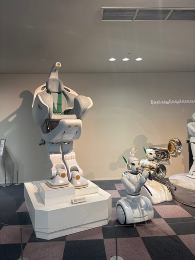 The image captures an exhibition featuring robots in Nagoya, Japan, likely at the Miraikan (National Museum of Emerging Science and Innovation) given the nature of the exhibits. The central focus is a large, white, chair-like robot with a grey seat and green restraints, resembling a high-tech, possibly interactive, vehicle. To its right, two smaller, humanoid robots are prominently displayed. One robot, positioned closer to the viewer, is playing a brass instrument, likely a trombone or tuba, with its arms extended. Another robot is partially visible behind it, also with brass instruments. The robots are situated on a checkered floor with a pattern of dark gray and pinkish-purple squares. In the background, a light beige wall displays the text "Today for tomorrow" in a stylized font. Several white pedestals or display bases support the robots. A sign that reads "Do not touch" in Japanese and English is visible on the base of the large robot. The overall environment appears to be an indoor exhibition space, well-lit with overhead lighting.