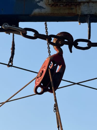 The image depicts a rusty, weathered pulley system against a clear, pale blue sky. The system is connected to a thick, dark metal support structure with visible paint chipping and rust. A heavy chain is connected to the support and extends down to a shackle. The pulley itself is painted red, though the paint is faded and worn. A rope is threaded through the pulley, extending to the right. Additional ropes and chains are also present. The location in the city of Emona, Bulgaria, is not discernible from the image.