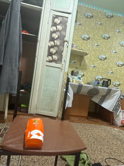 The image depicts an interior scene, possibly a residential room, in Corlăteni, Moldova. In the foreground, an orange box, possibly food packaging, rests on a brown surface. To the left, a dark grey towel hangs from a wooden structure, partially obscuring a closet. The closet contains shelves with various items, including clothing and containers. A tall, off-white door with a decorative panel featuring floral motifs stands in the center. To the right, a table is covered with a white tablecloth adorned with a black geometric pattern. On the table, several bottles, a black electric kettle, and a white bag are visible. The wall in the background is decorated with yellow wallpaper featuring a stone pattern and circular motifs of houses. Blue string lights are visible along the top of the wall. The floor is covered with a patterned carpet and a section of wooden flooring. A green object, possibly footwear, is visible on the carpet in the lower right corner. The overall impression is of a cluttered domestic space.