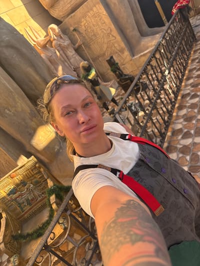 A low-angle selfie depicts a person wearing a white shirt, a grey vest with purple buttons and red suspenders, and having blonde hair and tattoos on their arm. They are indoors, standing near an ancient Egyptian display. The background features Egyptian artifacts, including hieroglyphs and sculptures of pharaohs and deities. The location is likely an exhibit or attraction related to ancient Egypt in Dahab, Egypt.