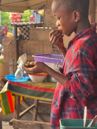 The image, rotated 90 degrees clockwise, shows a young person in profile, likely a boy, eating something with his hands in what appears to be an outdoor market or street stall setting.
The person is positioned in the left half of the frame, facing towards the right. They have short, dark hair and are wearing a red and dark blue plaid long-sleeved shirt. Their right hand is raised to their mouth, and they are in the process of taking a bite of food. Their left hand holds a small, crumpled wrapper or package, from which the food is being consumed.
In the mid-ground, behind the person, a purple plastic crate or basket is visible. To the left and slightly behind the person, various packaged goods in brightly colored wrappers (appearing in shades of blue, green, and red) are stacked or displayed on wooden shelves or a stall. Part of a dark-colored umbrella or canopy is visible on the far left side. The surface beneath these items has a black and white checkered pattern, resembling a checkered cloth or tile.
Further into the mid-ground and extending to the right side of the frame, additional wooden structures suggest market stalls or a framework. On the right side, a colorful striped fabric, featuring prominent green, red, and yellow stripes, is draped over an unseen object. Near this fabric, a white bowl with an orange rim and a blue plastic lid or container are visible. In the upper right corner of the frame, a green container holds several silver utensils, including forks and spoons.
The scene conveys a daytime setting with natural light, suggesting a casual, functional environment, likely a marketplace where goods are sold and food is readily available. The focus is on the person enjoying their meal amidst the backdrop of a bustling, open-air commercial area.
