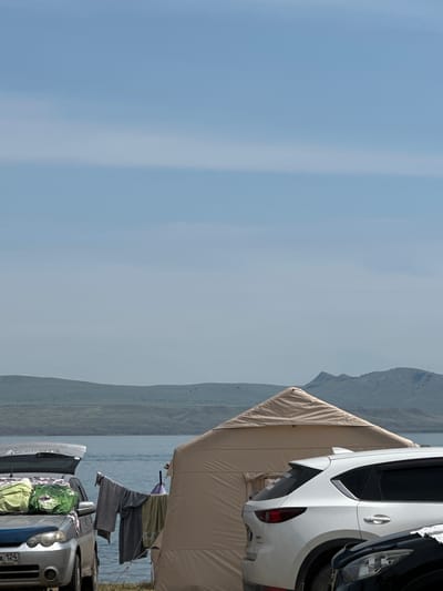 The image depicts a campsite near a body of water, possibly a lake or river, with hills in the background. Two cars are parked, one silver and one white. The silver car has its trunk open and several bags are piled on top of it. Clothes are hanging on a line strung between the silver car and a large tan tent. The white car is parked next to the tan tent, and the front of a black car is visible in the lower right corner of the image. The sky is clear with some faint clouds. The scene suggests a recreational or camping activity. The specific location of Колодезный, Russia, is not explicitly visible or indicated in the image.