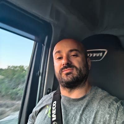 A man with a bald head and a beard is taking a selfie from inside a vehicle. He is wearing a grey t-shirt with a black strap across his chest that has white text on it. The man is looking at the camera with a slight smile. The background shows a window with a blurred view of trees and the sky. The interior of the vehicle is dark, with a seat visible behind the man. The scene appears to be a daylight setting, possibly during a drive. The image does not provide enough information to determine the specific location within Riba-roja de Túria, Spain.
