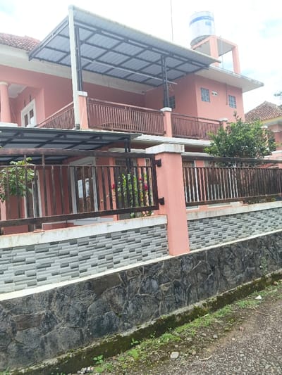 The image displays the exterior of a two-story residential building situated in Pô Kô, Vietnam. The building is painted a uniform salmon pink. A prominent corrugated metal canopy, supported by black metal framework, extends over the second-story balcony and partially covers the ground floor. The second story features a balcony with dark brown wooden railings. A white and blue "Penguin" brand water tank is mounted on an elevated salmon pink support structure on the roof's right side.

The property is fronted by a dark brown metal fence, similar in style to the balcony railings, with a salmon pink concrete pillar serving as a gatepost. Behind the fence, red flowering plants and a green bush are visible. A retaining wall below the fence comprises two distinct sections: an upper layer of grey and white horizontally patterned tiles, and a lower base constructed from dark, irregularly shaped natural stones. A dirt and gravel path, interspersed with sparse green vegetation and moss, runs along the base of the stone wall in the foreground. White-framed windows and doors are visible on both levels of the building. A white garment hanger is suspended from the underside of the canopy on the second floor. No individuals or animals are present in the scene.