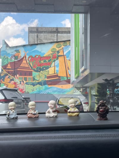 The image shows a view from inside a vehicle, looking through the windshield at a mural on a building. The mural depicts a traditional Indonesian scene with houses, boats, and people, and the text "Welcome to Dumai" is prominently displayed. In the foreground, on the dashboard of the vehicle, are several small figurines, including a row of five meditating monks and a laughing Buddha statue. Behind the figurines and through the windshield, cars are visible parked in front of the muraled building and a modern building with green accents. The sky above is blue with scattered clouds. The scene appears to be in Dumai City, Indonesia, as indicated by the mural.