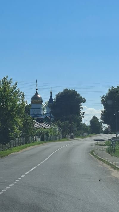 An asphalt road curves to the right under a clear blue sky. On the left side of the road, a wooden fence lines a grassy area. Behind the fence, a cluster of trees partially obscures a white building with a golden dome and a blue spire, likely a church. Further down the road, on the right side, a speed limit sign indicating 40 is visible. The environment appears to be a rural or semi-rural setting in Хажоўскі сельскі Савет, Belarus.