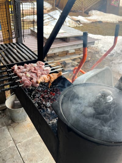 The image depicts a cooking scene, likely outdoors. Skewers of meat and poultry are arranged on a grill over hot coals, with a large black pot positioned nearby. An orange wheelbarrow sits in the background. The location is potentially Bol'shakino, Russia.