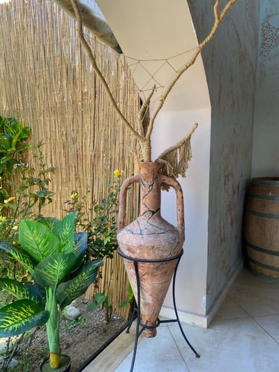 This media file shows an outdoor scene with decorative elements. The main subject is an antique-looking terracotta amphora, supported by a black metal stand. The amphora is adorned with frayed rope that resembles hair or a headdress, and appears cracked and weathered.

The amphora is placed in front of a bamboo-reed screen. To the left, a white, curved structure, possibly a wall or awning, is visible, with thick ropes and netting attached to it, creating a decorative canopy. In the upper background, a wooden barrel is partially visible.

In the foreground, lush green plants with large, variegated leaves are growing, some of which appear to be in planters. A small yellow flower is also visible near the amphora, blooming amongst the greenery. The ground beneath the plants is a mix of soil, small stones, and gravel.

The lighting suggests it is daytime, with soft sunlight filtering through. The overall impression is one of a rustic, Mediterranean-inspired decorative setting, possibly in a garden or courtyard. The location context of Kyrenia, Northern Cyprus/Cyprus, aligns with the visual elements of sun, plants, and architectural style. No people are visible in the image. No text is discernible.