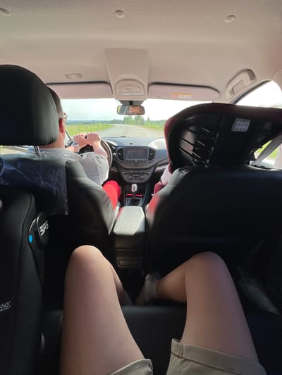 A view from the back seat of a car traveling on a road in Sidorovy Gory, Russia. The driver is visible, wearing a light-colored shirt, red pants, and a watch. Their hands are on the steering wheel. A child car seat is in the back seat, and an individual's leg is visible in the back passenger seat. The car's dashboard, console, and rearview mirror are visible, as well as the external environment seen through the windshield.