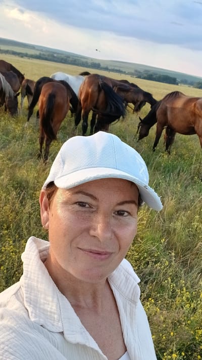 A woman wearing a white cap and white collared shirt takes a selfie in a grassy field with a herd of horses in the background. The sky is cloudy with hints of sunset colors. The location is Nadezhdenka, Russia.
