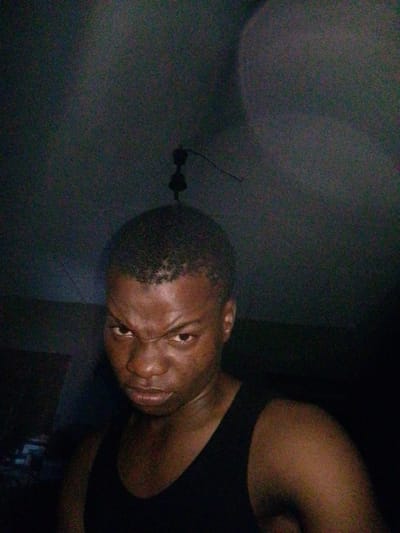 The image is a close-up, dimly lit selfie of a young Black male. He is wearing a black tank top and has a stern expression with his eyebrows furrowed. The background is dark and appears to be an indoor setting, possibly a ceiling fan is visible above his head. The quality of the image is grainy, suggesting it was taken with a low-light setting or an older camera. There is no discernible information in the image that indicates a specific location within Eleweran, Nigeria, or any broader urban context.