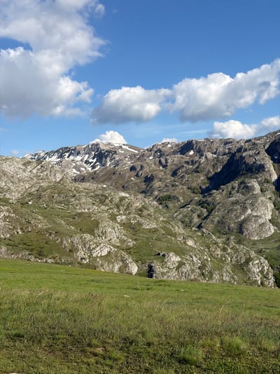 The image shows a mountain landscape in Boričje, Montenegro. The left side of the image features a blue sky with white, fluffy clouds. The majority of the image is dominated by a large, rocky mountain with patches of snow, especially on its upper slopes. The lower part of the image features a green, grassy meadow that borders the mountain.