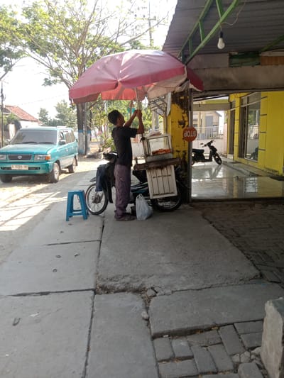 A man is adjusting a red and white umbrella on a street in Kemamang, Indonesia. He is standing next to a motorcycle with a cart attached, which is likely used for selling goods. A blue plastic stool is placed on the sidewalk in front of the motorcycle. To the left, a teal colored SUV is parked on the street. The background shows a building with a yellow facade and a covered outdoor area with a hanging light bulb and a STOP sign. Trees are visible in the upper left corner of the image, suggesting an outdoor, possibly commercial area. The ground is a mix of concrete and pavement.