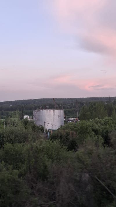 The image shows an outdoor scene at dusk with a pale pink and purple sky. The landscape is dominated by dense green trees on the right side, obscuring the background. In the center-left of the image, a large, cylindrical, metallic structure is visible, possibly a storage tank or part of an industrial facility. A crane is positioned near this structure, suggesting ongoing construction or maintenance work. In the background, beyond the trees, a dark, forested hillside stretches across the horizon. The overall impression is of an industrial area encroached upon by nature. The time of day is indicated by the soft, diffused light and the colors in the sky, suggesting twilight. There is no visible text in the image. The location context is Achinsk, Russia.