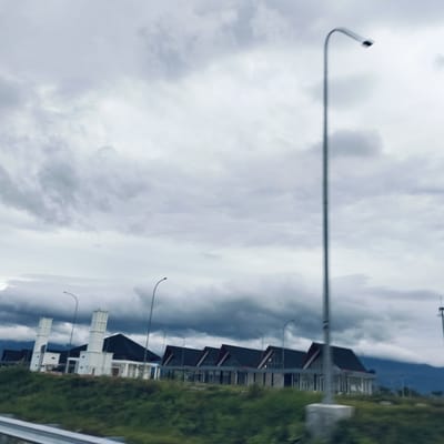 The image depicts an overcast and cloudy day in Cotkareung, Indonesia, showcasing a series of buildings with distinctive architecture. The scene is captured from a slightly elevated perspective, suggesting it was taken from a moving vehicle on a road or highway.
In the mid-ground, a row of structures features prominent dark, steep-sloping roofs and mostly white facades. On the left, two tall, white, tower-like elements, possibly minarets or decorative architectural features, rise beside a main building. To their right, several similar buildings are arranged sequentially, each with a dark, gabled roof and large windowed sections. These buildings appear new or recently constructed, possibly part of a rest area or a commercial development. Several streetlights are visible throughout the mid-ground, illuminating what appears to be a parking area or access road in front of the buildings.
The upper portion of the frame is dominated by a heavy, grey, and dark cloudy sky, creating a dim and moody atmosphere. The clouds are dense and layered, hinting at impending rain or a generally gloomy day. In the far background, faint outlines of mountains can be seen beneath the thick cloud cover.
The foreground is blurred, showing a green, grassy embankment and a metallic guardrail, reinforcing the impression of a moving vantage point. A tall, slender streetlight pole stands prominently in the right foreground, partially obscuring the view of the buildings. There is no visible human activity or text in the image. The overall impression is one of quiet solitude under a dramatic sky.