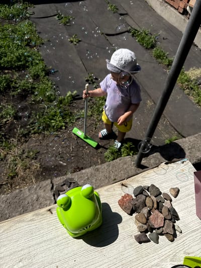 In a rural setting near Zykova, Russia, a young child wearing a light-colored hat and a purple t-shirt is playing with a bubble wand in a yard. The ground is covered with dark fabric and patches of green grass. The child is holding a green broom-like bubble wand, and numerous iridescent bubbles are floating in the air around them. To the foreground, on a weathered wooden surface, sits a bright green toy frog and a pile of small, varied-colored rocks. The scene is illuminated by bright sunlight, casting shadows and highlighting the floating bubbles.