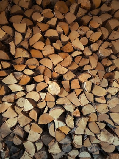 The image displays a dense pile of chopped firewood. The wood pieces vary in size and shape, with many showing the characteristic circular or semi-circular cross-sections of logs. The color of the wood ranges from light tan to darker brown, with some pieces exhibiting reddish hues. The texture appears rough and natural, with visible grain patterns and bark on some sections. The firewood is stacked tightly, creating a uniform, textured background. The lighting suggests an indoor or shaded outdoor environment, casting subtle shadows that highlight the three-dimensional nature of the woodpile. There are no people or animals visible in the image, and no specific location within Vecumnieki, Latvia is discernible from the visual information.