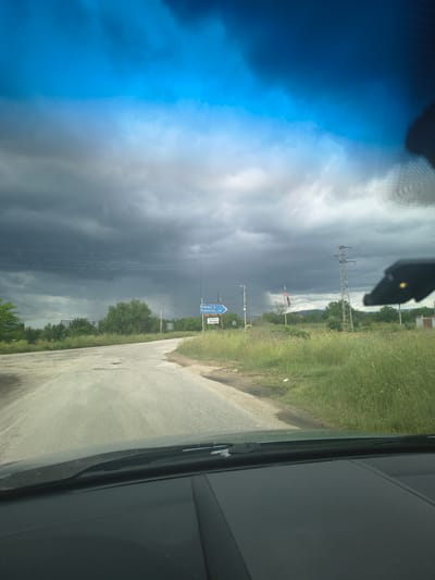 The image is taken from inside a vehicle, likely the driver's seat, looking out onto a rural road in Padarsko, Bulgaria. Overcast skies, indicative of an approaching storm, dominate the left side of the frame. A road stretches along the right side, with vegetation growing along its edge. A signpost displaying directions indicates distances to nearby towns, specifically "Борци 4" (Borci 4) and "Брезово 18" (Brezovo 18). Power lines run alongside the road, and a Bulgarian flag is visible in the distance.