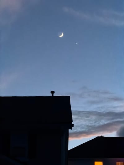The image is a vertical, eye-level shot of the sky at dusk, with the silhouettes of two houses in the foreground. The sky is a deep blue, transitioning to lighter hues near the horizon where clouds are visible, tinged with pink and orange. High in the sky, a crescent moon is visible to the left, and a small, bright star-like object, likely a planet such as Venus, is located to the right of the moon. Another, fainter celestial body is visible in the upper right quadrant of the sky. The house on the left is a dark silhouette with a visible vent pipe on its roof. The house on the right is partially visible, with a section of roof silhouetted against the sky. One window on the lower right side of the image emits a warm, orange glow. The scene appears to be in a residential area, typical of Buford, Georgia, United States, given the presence of suburban housing. There are no people visible in the image.