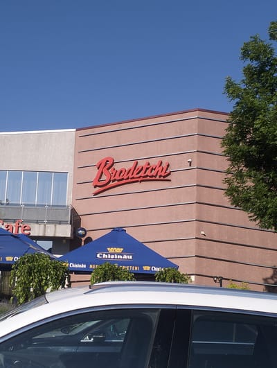 The image shows a building with a prominent red sign that reads "Brodetchi". The building has a modern design with horizontal lines on its facade. To the left of the "Brodetchi" sign, a section of the building has a lighter-colored facade and large windows. In front of the building, there are blue umbrellas, suggesting an outdoor seating area. A white car is partially visible in the foreground. A tree with green foliage is located to the right of the building. The sky is clear and blue. The location is specified as Șoldănești, Moldova.