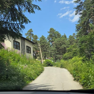The image shows a paved road winding through a densely forested area with a clear blue sky and scattered white clouds. On the left side of the road, a building with a light-colored facade and stone accents is visible, partially obscured by lush green vegetation and wildflowers. The building has two dark-framed windows. A wooden utility pole with wires runs along the side of the road, disappearing into the trees behind. The road appears to be in the village of Atoluka, Bulgaria, given the characteristic architecture and natural setting. The overall scene depicts a rural or semi-rural environment on a sunny day.