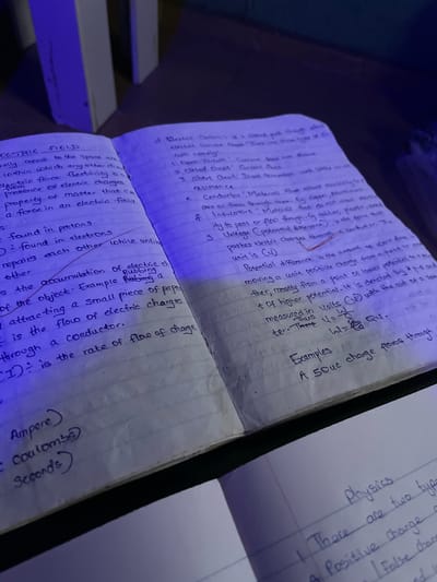 The image shows an open notebook with handwritten notes on physics. The notes are written in blue ink on lined paper. The notebook is lying on a dark, wooden surface, possibly a desk. To the left of the notebook, a portion of a white, vertical object is visible, which could be a wall or furniture. The lighting in the scene is dim and has a blueish tint, suggesting artificial light. The focus is on the notebook and its contents.
The notes on the left page are titled "ELECTRIC FIELD". They define electric field, electric force, and property of matter. There are also entries for "found in protons" and "found in electrons", as well as a definition related to the accumulation of electric charges and attracting small pieces of paper. The bottom of the left page contains notes on "Ampere", "Coulombs", and "Seconds".
The right page of the notebook has notes on "Electric Current", listing three types: "Open Circuit", "Closed Circuit", and "Short Circuit". It also defines "Resistance" and lists "Conductors" and "Insulators" with examples. Further down, there are notes on "Potential difference", defining it and its unit, and a formula related to it. The section "Examples" on the right page mentions "A 50uc charge flows through".
At the bottom right of the image, a separate page, possibly from another notebook or document, shows the word "Physics" and starts listing types of charge.
The location of Edopa, Nigeria is not mentioned or inferable from the image.