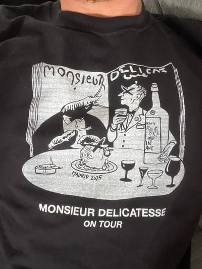 The image displays a black t-shirt featuring a detailed, cartoon-style graphic in white and light gray, possibly being worn by someone, with a portion of an arm visible on the left. The scene depicts a culinary or wine-tasting event.

**Subjects:** The graphic features two stylized male figures. A larger figure, dressed in what appears to be a uniform or suit, holds a glass and gestures towards the setting, possibly presenting or serving. Beside him, a large wine bottle labeled "ALSACE" and "VIN BLANC" is prominent, surrounded by various empty and filled wine glasses. Below, a smaller figure is seated at a table, appearing to eat with a fork near his mouth. The table is laden with food items, including a plate with what looks like seafood, a large shrimp or prawn, a small bowl with an anchor motif, and another small dish.

**Activity or event taking place:** The figures are engaged in dining and drinking, suggesting a gourmet experience, wine tasting, or a celebratory meal. The "ON TOUR" text implies a series of events.

**Notable details:** The artwork has a distinct hand-drawn, illustrative quality, using a limited color palette of black and white/gray. The text "MADRID 2025" is visible near the smaller figure, indicating a specific future event location and year. No specific time of day, weather, or discernible emotions are depicted.

**Visible text:** Key phrases include "MONSIEUR DELICATESSE" (written vertically on the left side of the graphic and on the right side, followed by "ON TOUR"), "ALSACE" and "VIN BLANC" on the wine bottle label, and "MADRID 2025."
