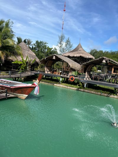 The image depicts a picturesque scene in Tambon Pa Kae Bo Hin, Thailand, featuring a tranquil turquoise body of water. In the foreground, a traditional longtail boat, painted a deep red, is moored to a wooden dock. The boat is adorned with a colorful fabric tied to its bow. Across the water, a series of thatched-roof huts are situated on elevated wooden platforms, accessible by walkways. These structures appear to be open-air dining or seating areas, with tables and chairs visible within some of them. One of the huts has a bright orange life preserver hanging on its side. A small fountain in the water sprays water upwards, creating a pleasant visual effect. Palm trees and lush greenery surround the huts, adding to the tropical ambiance. In the background, a tall communication tower rises against a bright blue sky dotted with wispy clouds. The scene suggests a peaceful, resort-like setting, ideal for relaxation and enjoying the natural beauty of the region.