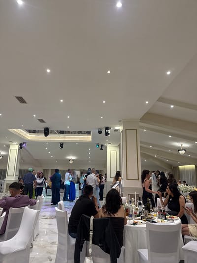The image shows an interior view of a large hall with a high ceiling. There are people sitting at tables covered with white cloths and white chairs, and more people are standing and conversing near a dance floor. Some of the people at the tables are looking at their phones or interacting with the table's contents. The dance floor appears to have a multicolored light pattern projected onto it. The ceiling contains multiple spotlights and ornate ceiling fixtures. The walls are white with decorative moldings. The precise location in Veselinovo, Bulgaria cannot be determined from this image alone.