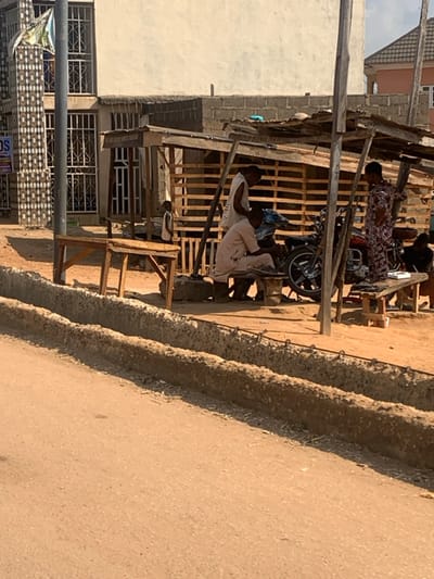 This image captures an outdoor street scene in Egbedi, Nigeria, under daylight. In the foreground, a reddish-brown dirt road is present, bordered on the right by two parallel, unfinished concrete structures, possibly for drainage, which feature exposed rebar.
Midground features a makeshift wooden shelter with a corrugated roof, supported by poles. Beneath this shelter, a dark-colored motorcycle is centrally positioned. A man in light-colored traditional attire is seated on a low wooden bench in front of the motorcycle, appearing to be engaged with it. Another man, wearing a white sleeveless top, stands directly behind the seated man. To their right, a woman in a multi-colored patterned dress stands, holding an object in her right hand. A fourth individual is partially visible, seated on a bench to the far right. Further left, adjacent to the shelter, a child in a white vest is partially visible behind an empty wooden table.
The background on the left features a multi-story building with a light-colored facade, distinguished by a pillar with decorative dark and light-colored tiles and multiple windows fitted with metal security grates. Other less defined residential or commercial buildings are visible further in the background.