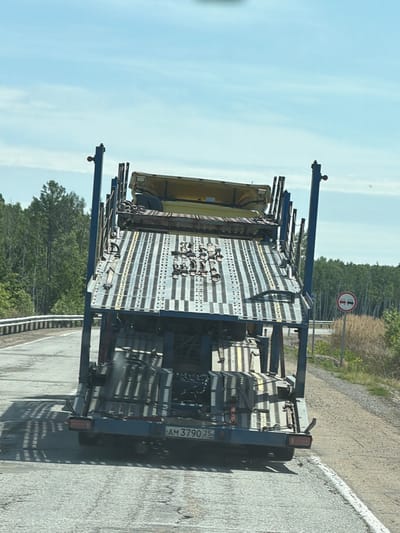 The image shows the rear view of a blue car carrier trailer truck driving on a paved road in a rural area. The truck is loaded with what appears to be car parts or equipment. The road is cracked and worn, with a white line marking the edge. To the right of the road, there is dry grass and a line of trees in the distance. A circular traffic sign with two cars in red and white is visible on a pole to the right. The sky is blue with some wispy clouds. The license plate of the truck is visible and reads "AM 3790 25". The environment suggests a location in a sparsely populated area, likely on a highway or provincial road. There are no people visible in the image. The location is stated as Аур, Russia, which is a town in the Republic of Buryatia.