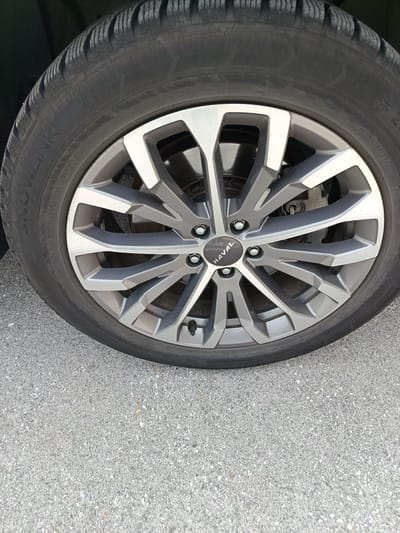 A close-up, eye-level, outdoor, daytime, vertical, full shot shows the front right wheel of a Haval SUV parked on a grey asphalt road. The tire is a black, SnowLink brand tire, and the rim is a multi-spoke alloy wheel with silver and grey accents. The Haval logo is visible in the center of the wheel. The background is blurred, showing the dark body of the car and a grey concrete wall. There are no people or animals visible in the image. The image was taken in Hallein, Austria.