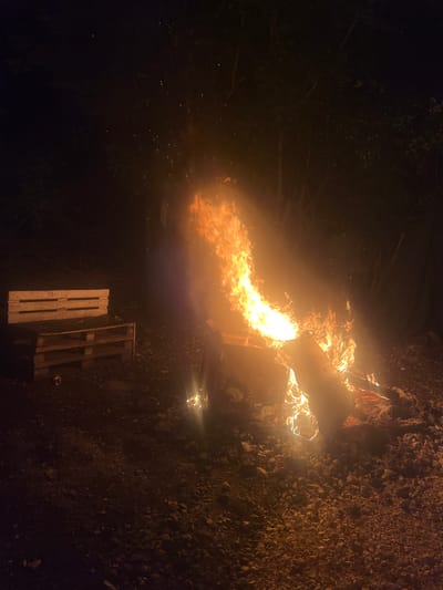 The image depicts a large, vibrant bonfire or controlled burn taking place at night in Pobeda, Bulgaria. The scene is dominated by intense, bright orange and yellow flames erupting from a pile of burning materials, including what appears to be a large metal object or container, and wooden debris. Smoke and numerous glowing sparks are visible rising into the dark night sky, illuminated by the fire's powerful glow.
The ground surrounding the fire, particularly to the right, is covered with coarse gravel and small rocks, clearly visible under the firelight. On the ground below the main flames, two wooden pallets are resting, largely intact, with another pallet seemingly consumed by the fire itself. The left side of the frame reveals dark, silhouetted foliage, suggesting trees or dense bushes, indicating an outdoor, possibly rural or undeveloped setting. A small, bright artificial light source, possibly a flashlight or lantern, is visible in the lower right corner, casting a localized glow on the ground.
The activity captured is a significant burning event, likely a bonfire or the disposal of materials by fire. The time of day is clearly night, with the fire being the primary light source. There are no people visible, and no discernible text in the image.