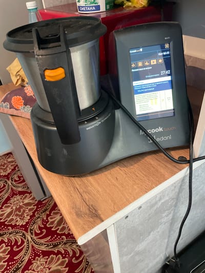 The image shows a modern kitchen appliance, a multicooker, placed on a wooden countertop. The multicooker has a silver-colored bowl and a black lid with a prominent orange handle release button. A digital display on the front of the appliance shows a recipe for "Rice porridge with milk," with a remaining time of 27:42. The screen also indicates "Het Wi-Fi," suggesting it is connected to a wireless network. The brand name "Mcook touch" and "Tedani" are visible on the appliance. To the left of the multicooker, a partially visible bottle of water with a white cap and a red object are present. The appliance is situated on a wooden surface, with a red and beige patterned rug visible on the floor in the foreground. The background shows a glimpse of a kitchen setting. The overall scene depicts a domestic environment, likely a kitchen, where food preparation is in progress. The image does not provide any information or visual cues related to the city of Bujumbura, Kyrgyzstan.