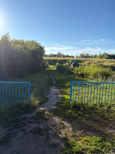 The image depicts an outdoor scene on a sunny day, likely in Pobedinka, Russia, given the context provided. In the foreground, a blue metal fence with vertical bars is visible on the left and right sides of the frame, with an opening in the center. Beyond the fence, a dirt path winds its way through grassy terrain, leading towards a small, dark-roofed wooden building with a white window, possibly a chapel or small house. This building is situated amidst lush green bushes and trees, on a gentle slope. The sky above is a clear blue with a few wispy white clouds. In the distance, power lines on poles are visible against the horizon, which is lined with trees and vegetation. The overall impression is of a rural or semi-rural landscape.