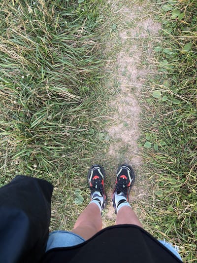 A first-person perspective shot captures a person standing on a dirt path surrounded by grass. The person is wearing black athletic shoes with red and white accents, and light-colored socks with black stripes. Denim shorts and a black skirt or shorts are visible. The scene appears to be outdoors in a natural setting, possibly a park or trail, with the sun casting a light shadow on the path, suggesting it is daytime. The grass is green and somewhat overgrown, with scattered small white flowers. The ground is a mix of dirt and grass. The overall impression is casual and active, likely taken during a walk or outdoor activity. No text is visible in the image. The location context provided is Mar'inskoe, Russia.