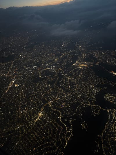 An aerial, oblique night view depicts the illuminated urban landscape of Atizapán de Zaragoza, Mexico. The scene is characterized by a vast, intricate network of golden and white artificial lights delineating residential areas, commercial zones, and arterial roads across the dark terrain. A prominent, irregular dark expanse, identified as the Presa Madín reservoir, occupies the middle-right and bottom-right portions of the frame, bordered by the illuminated city. Above the urban sprawl, dark, layered cloud formations partially obscure the night sky. A sliver of twilight-colored sky, exhibiting hues of orange and pink, is visible above the highest cloud layer at the top of the image, indicating either late dusk or early dawn. No specific individuals, actions, or interactions are discernible from this elevated perspective. The environment consists of the illuminated city, the dark reservoir, and an overcast sky.