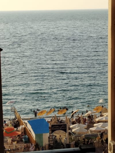 A view from a balcony overlooking the Mediterranean Sea, located at the Regency Beach in Alexandria, Egypt (latitude: 31.273, longitude: 30.001). The sea is calm and blue with a light breeze, with some people swimming and sunbathing in the distance. On the beach, there are multiple yellow and white beach umbrellas, along with some chairs and tables. People are scattered around the beach, enjoying the sun and the sea. There is a small blue building in the foreground, with a sign that reads "Regency Beach" in Arabic and English. The view is from a building, with a brown wall to the right and a large window looking out over the scene. The sun is setting in the distance, casting a golden hue over the scene.