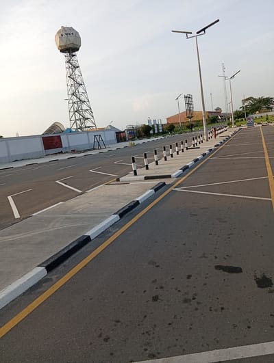The image depicts a paved area in Omagwa, Nigeria, likely a parking lot. The pavement is dark asphalt with yellow and white painted lines designating parking spaces and traffic flow. A raised concrete median runs along the edge of the pavement, marked with alternating black and white paint. A tall, cylindrical NIMET radar tower is visible in the background, along with other structures and utility poles. The sky is light and hazy. The scene appears to be deserted except for what appear to be two figures in the background. The ground shows signs of wear and some staining.