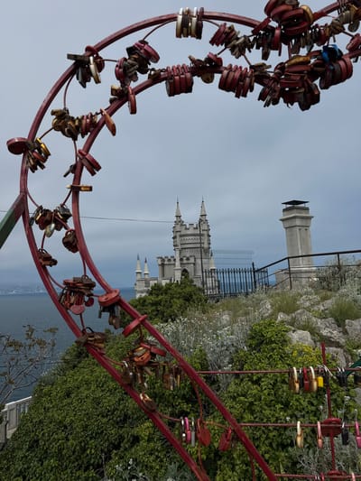 The image shows a red metal structure adorned with numerous padlocks, likely a "love lock" installation. In the background, the Swallow's Nest castle can be seen perched on a cliff overlooking the sea. The vegetation is lush and green, suggesting a coastal environment. The image appears to have been taken in the Гаспринский поселковый совет in Ukraine.