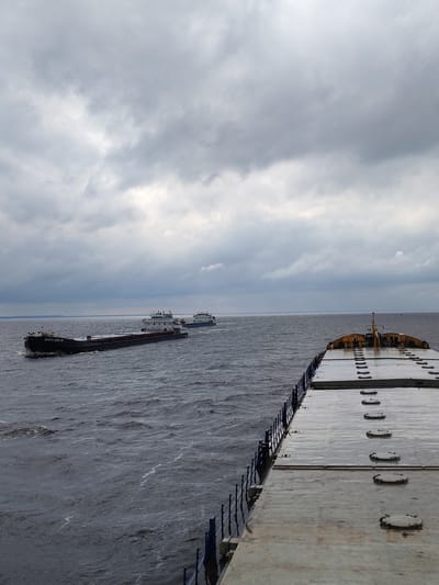 This image captures a wide shot of a body of water, likely a river or canal, under an overcast sky. In the foreground, the deck of a large vessel occupies the right half of the frame, showing a metallic surface with numerous circular hatches and a blue railing. The rest of the image is dominated by the water and the sky.

Further out on the water, two cargo ships are visible. The closer one, on the left, is a long, flat-bottomed barge identified by the Cyrillic text "ВОЛГО-ДОН 101" on its hull. It is moving forward, creating a wake. Behind it, a second, smaller vessel is also proceeding in the same direction. The sky is filled with heavy, grey clouds, suggesting a cloudy or potentially stormy day. The water is dark and appears somewhat choppy, with small waves. The horizon is a faint line separating the water from the distant shore. The overall atmosphere is somber, conveyed by the muted colors and the dramatic sky. The scene appears to be set in Russia, possibly Novaya Svobodka, based on the provided location context, and the ships' names suggest they are part of a Russian fleet. The time of day is not clearly discernible due to the cloudy conditions, but it appears to be daytime. No people are visible in the image.